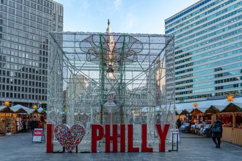 A festive display with "I ❤️ PHILLY" in front of a holiday market and city buildings in Philadelphia.