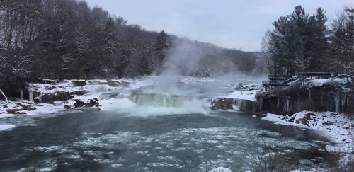 A snowy landscape featuring a waterfall with steam rising, surrounded by trees and icy riverbanks.