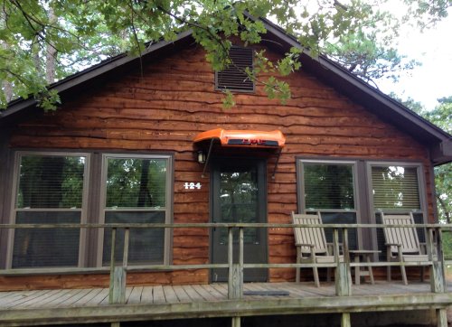 A rustic wooden cabin with a porch, featuring two chairs and large windows, surrounded by trees.
