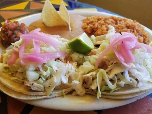 A plate of tacos topped with cabbage and pickled onions, served with rice, beans, salsa, and tortilla chips.
