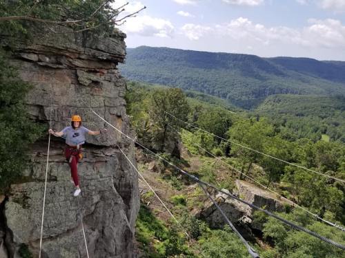A person in a helmet balances on a tightrope between cliffs, surrounded by lush green mountains and a blue sky.