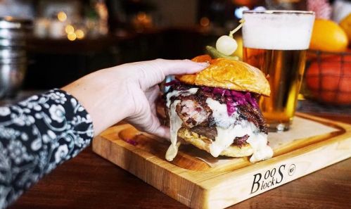 A hand holding a juicy burger with melted cheese and toppings, next to a glass of beer on a wooden board.