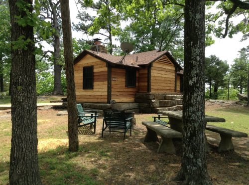 A rustic log cabin surrounded by trees, featuring a stone foundation, picnic table, and satellite dish.