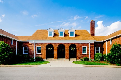 A brick building with a brown roof, arched entrance, and greenery under a blue sky.