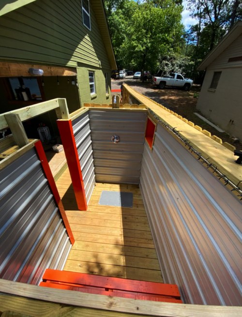 A wooden outdoor shower with metal walls, featuring a wooden floor and bright orange accents, surrounded by trees.