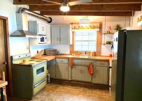 A cozy kitchen with wooden beams, green appliances, and a bright window adorned with colorful decorations.