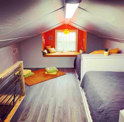 Cozy attic room with two beds, bright orange accents, a window seat, and soft seating on a wooden floor.