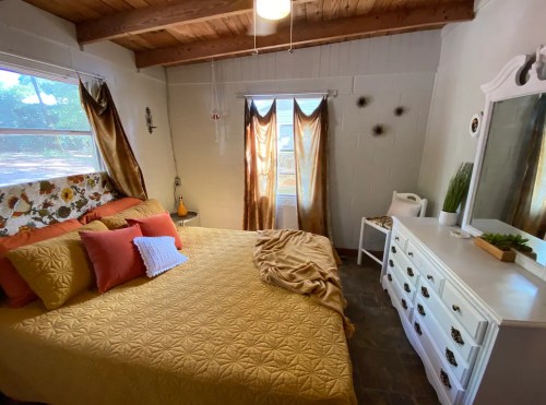 Cozy bedroom with a yellow bedspread, decorative pillows, and natural light from windows with sheer curtains.