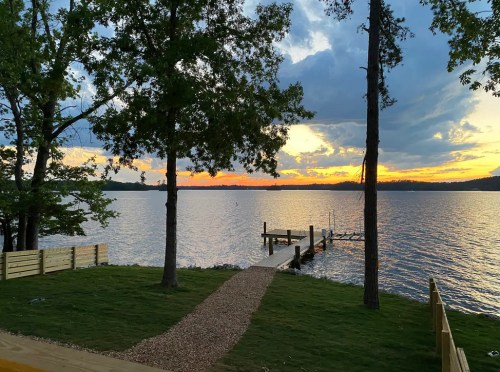 A serene lakeside view at sunset, featuring a dock, trees, and colorful clouds reflecting on the water.