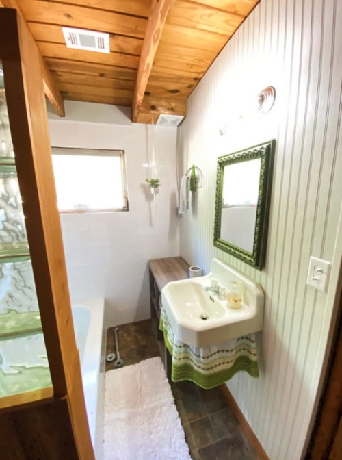 Cozy bathroom with wooden ceiling, white walls, a sink, mirror, and a bathtub, featuring green accents and a rug.