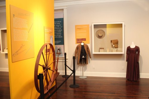 Exhibit featuring historical clothing and artifacts, including a spinning wheel and display cases with garments.