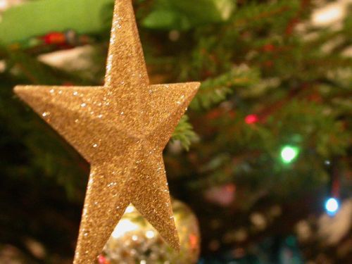 A glittery gold star ornament atop a Christmas tree, surrounded by green branches and colorful lights.
