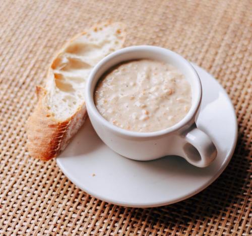 A small white cup filled with creamy soup, accompanied by a slice of crusty bread on a white plate.