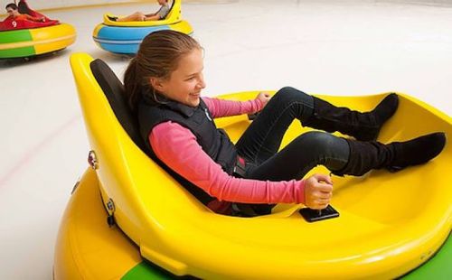 A girl smiles while riding a yellow bumper car on ice, with other colorful bumper cars in the background.
