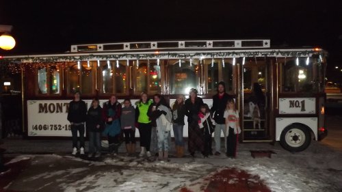 A group of people stands in front of a decorated trolley bus on a snowy night, smiling and posing for the photo.