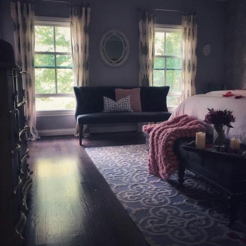 Cozy bedroom with a blue loveseat, large window, and soft decor, featuring a pink throw and candles on a dark table.