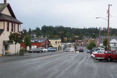 A quiet street lined with parked cars, leading to small buildings and trees in a coastal town under a cloudy sky.