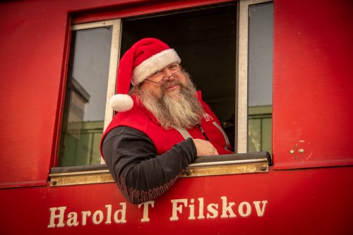 A man with a long beard and glasses wears a Santa hat, smiling from a window of a red train car.