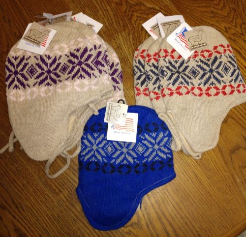 Three knitted hats in various colors with patterned designs, each with tags attached, displayed on a wooden surface.