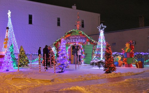Colorful holiday lights decorate a festive Santa's workshop scene with trees, gifts, and snowy surroundings.