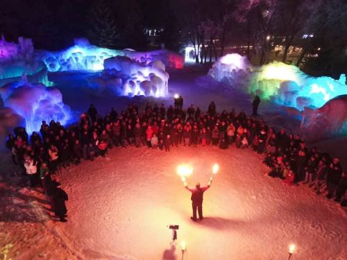 A crowd gathers around a performer juggling fire in a colorful, ice-lit winter landscape at night.