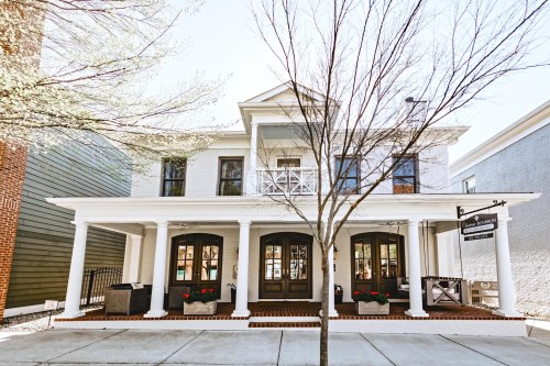 A charming two-story building with a white facade, large windows, and a porch, surrounded by trees and planters.