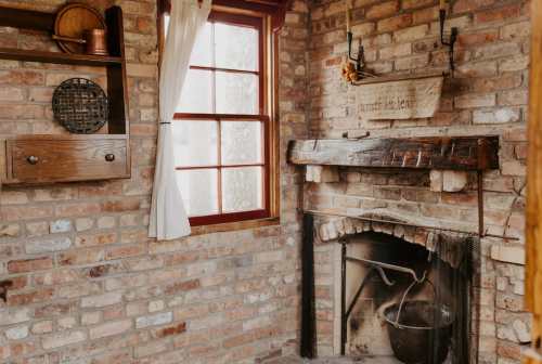 Cozy interior featuring a brick wall, wooden mantel, fireplace, and a window with white curtains.