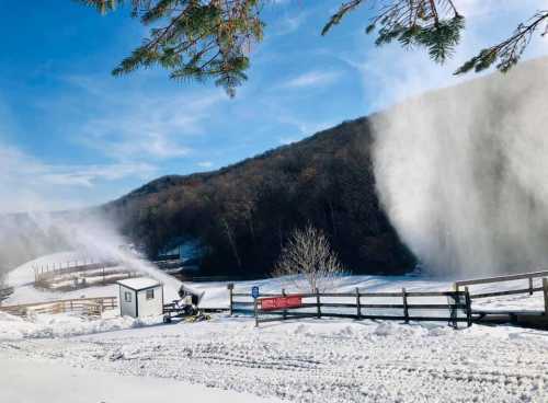 Snowmaking machines spray snow on a winter landscape, with a snowy hill and blue sky in the background.