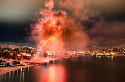 Colorful fireworks illuminate a snowy lakeside town at night, reflecting on the water below.