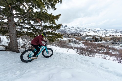 A person rides a fat bike down a snowy slope, overlooking a town surrounded by mountains.