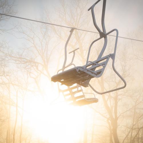 A ski lift chair hangs in the air against a misty background with sunlight filtering through bare trees.