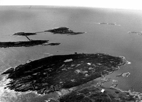 Aerial view of a rocky island surrounded by water, featuring scattered buildings and nearby smaller islands.