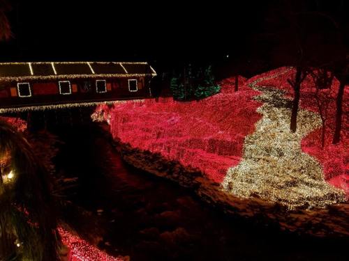 A covered bridge illuminated with colorful lights, surrounded by a glowing landscape of red and white lights at night.
