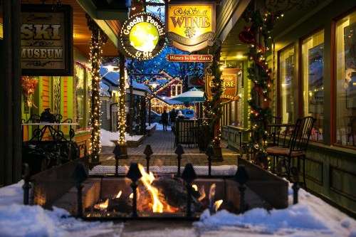 A cozy winter scene featuring a fire pit, festive lights, and shops along a snow-covered walkway.