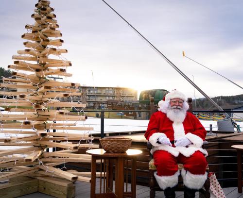 Santa Claus sits on a deck beside a wooden Christmas tree adorned with lights, with a scenic background at sunset.