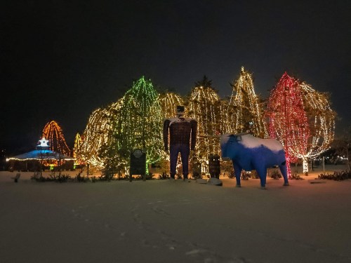 A snowy scene featuring colorful holiday lights on trees, a large figure, and a blue cow statue.