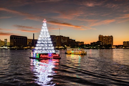A brightly lit Christmas tree floats on water at sunset, with city buildings in the background.