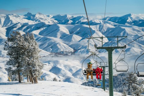 Two teddy bears sit on a ski lift, surrounded by snow-covered mountains and a clear blue sky.