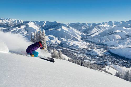 A skier carves through fresh snow on a mountain slope, with a stunning view of snow-covered peaks and a valley below.
