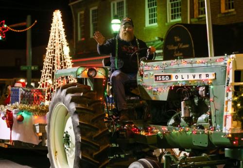 A man with a beard waves from a decorated tractor in a festive parade, surrounded by holiday lights and decorations.