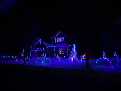 A house decorated with vibrant blue and purple Christmas lights, featuring illuminated trees and arches in the yard.