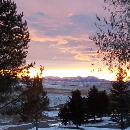 Sunset over snow-covered mountains, framed by trees, with a colorful sky in shades of orange and purple.