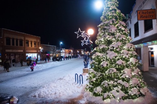 A snowy street at night, decorated with lights and a large Christmas tree, bustling with people enjoying the festive atmosphere.