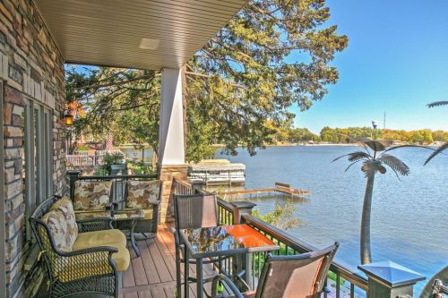 A cozy balcony with seating overlooks a serene lake, surrounded by trees and clear blue skies.