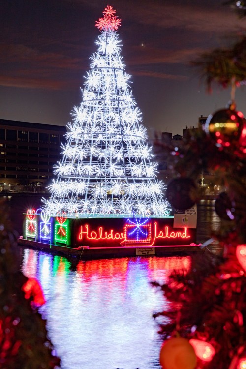 A brightly lit Christmas tree on water, with "Holiday Island" illuminated in colorful lights, surrounded by festive decorations.