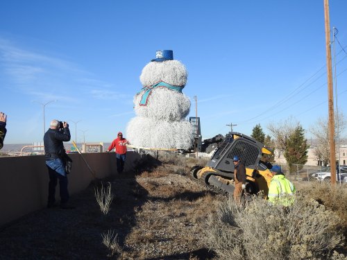 A large snowman made of white material stands on a roadside, while workers and onlookers gather nearby.