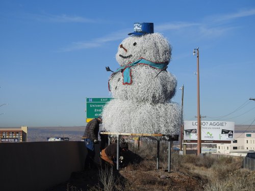 A large snowman made of wire sits on a platform by the highway, wearing a blue hat and scarf, with workers nearby.
