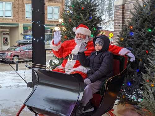 Santa Claus and a young boy wave from a festive sleigh, surrounded by Christmas trees and holiday lights.