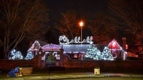 A festive building adorned with colorful Christmas lights, featuring a sleigh and reindeer on the roof, surrounded by trees.