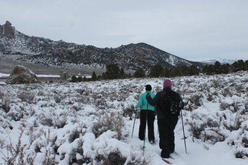 Two hikers trek through a snowy landscape, surrounded by mountains and sparse vegetation under a cloudy sky.